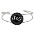 A silver-toned metal cuff bracelet featuring a round black disc with the word 'Joy' in white script, shown on a plain white background.