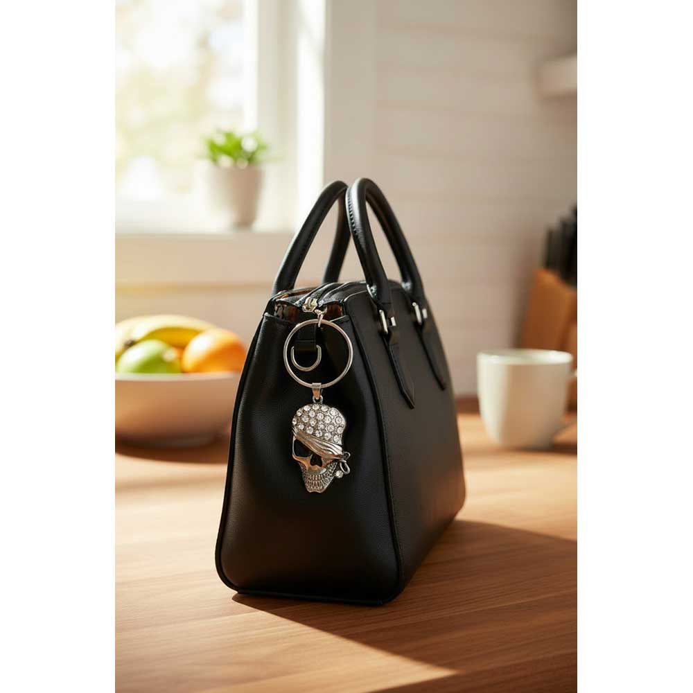 A black leather handbag with double top handles and a silver skull charm adorned with rhinestones, resting on a wooden table in a softly lit kitchen setting. Lilylin Designs