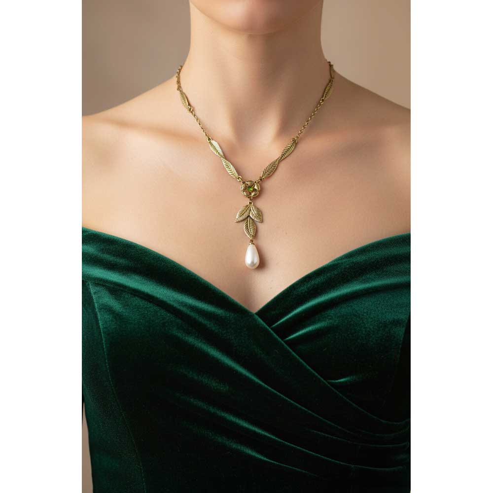 A gold necklace with a leaf and pearl pendant, shown on a model wearing a dark green velvet off-the-shoulder dress against a warm beige background. Lilylin Designs