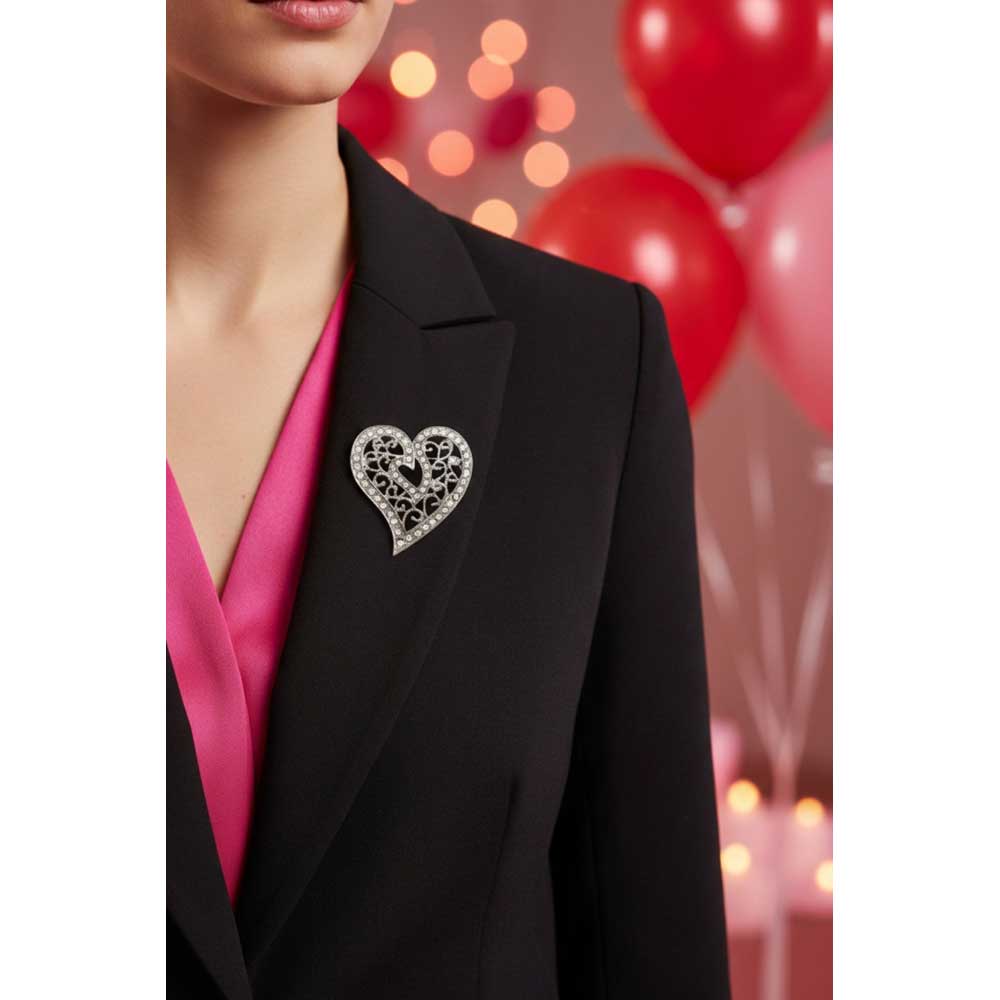 A silver heart-shaped brooch with intricate filigree detailing and small clear stones, worn on the lapel of a woman in a black blazer over a bright pink top. The background is a soft-focus pink scene with red balloons and warm bokeh lights. Lilylin Designs
