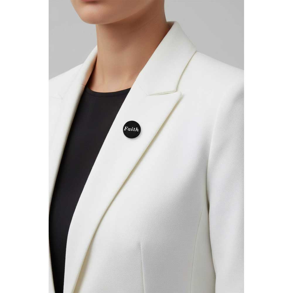 A close-up of a person wearing a white blazer over a black top, with a small round black lapel pin that reads 'Faith' on the chest. Lilylin Designs