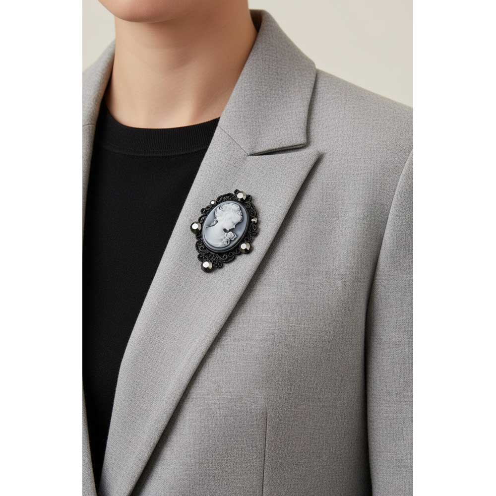 A close-up of a person wearing a light gray blazer over a black top, featuring a decorative black brooch with a white cameo-style face on the lapel. Lilylin Designs