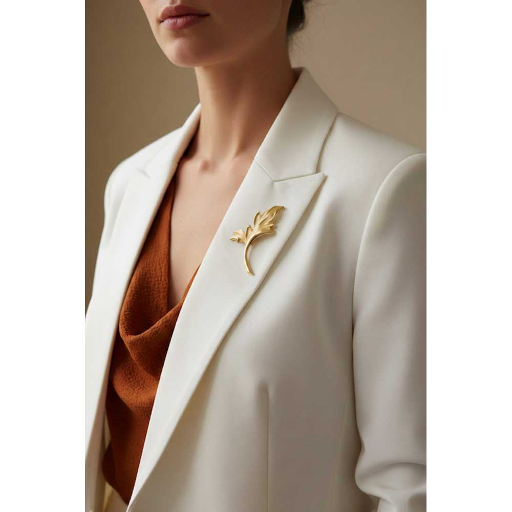 A close-up of a woman wearing a white blazer over a rust-colored top, featuring a gold leaf-shaped brooch pinned to the lapel.  Lilylin Designs