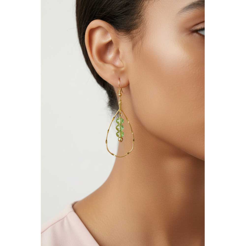 A close-up of a model's ear and neck wearing a gold teardrop hoop earring with a dangling green bead cluster. Lilylin Designs