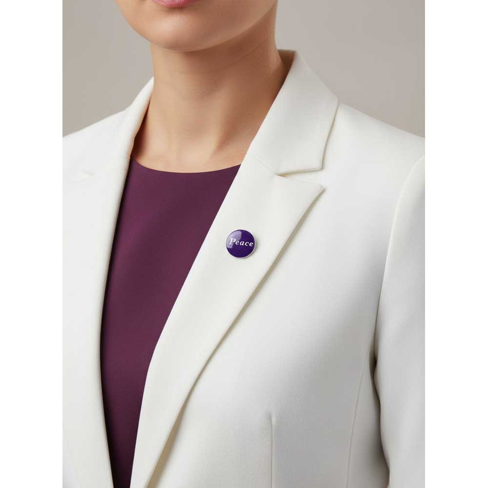 A close-up of a person wearing a white blazer over a deep purple top, with a small round purple lapel pin featuring the word 'Peace' pinned on the blazer. Lilylin Designs