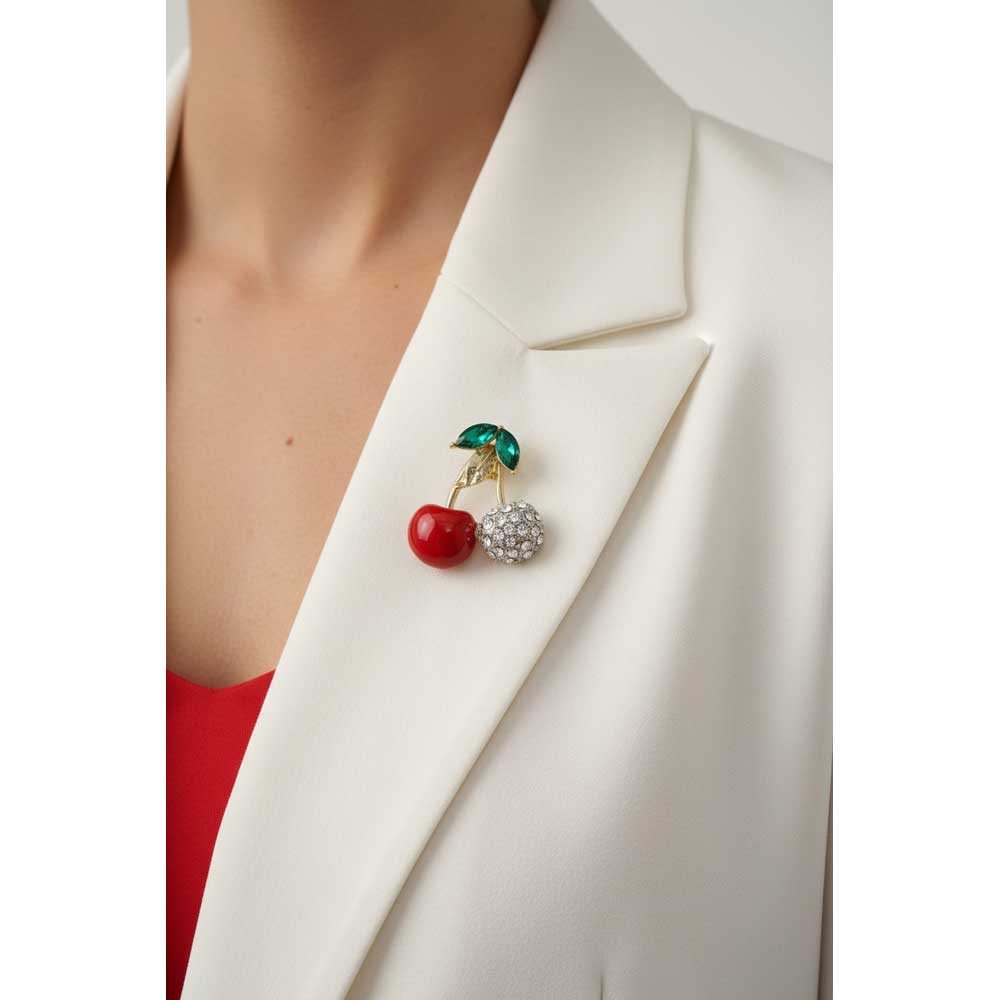 A close-up of a person wearing a white blazer and red top, with a cherry-themed brooch pinned to the lapel. The brooch features a red cherry, a silver-and-white rhinestone cherry, and green leaves with gold stems. Lilylin Designs