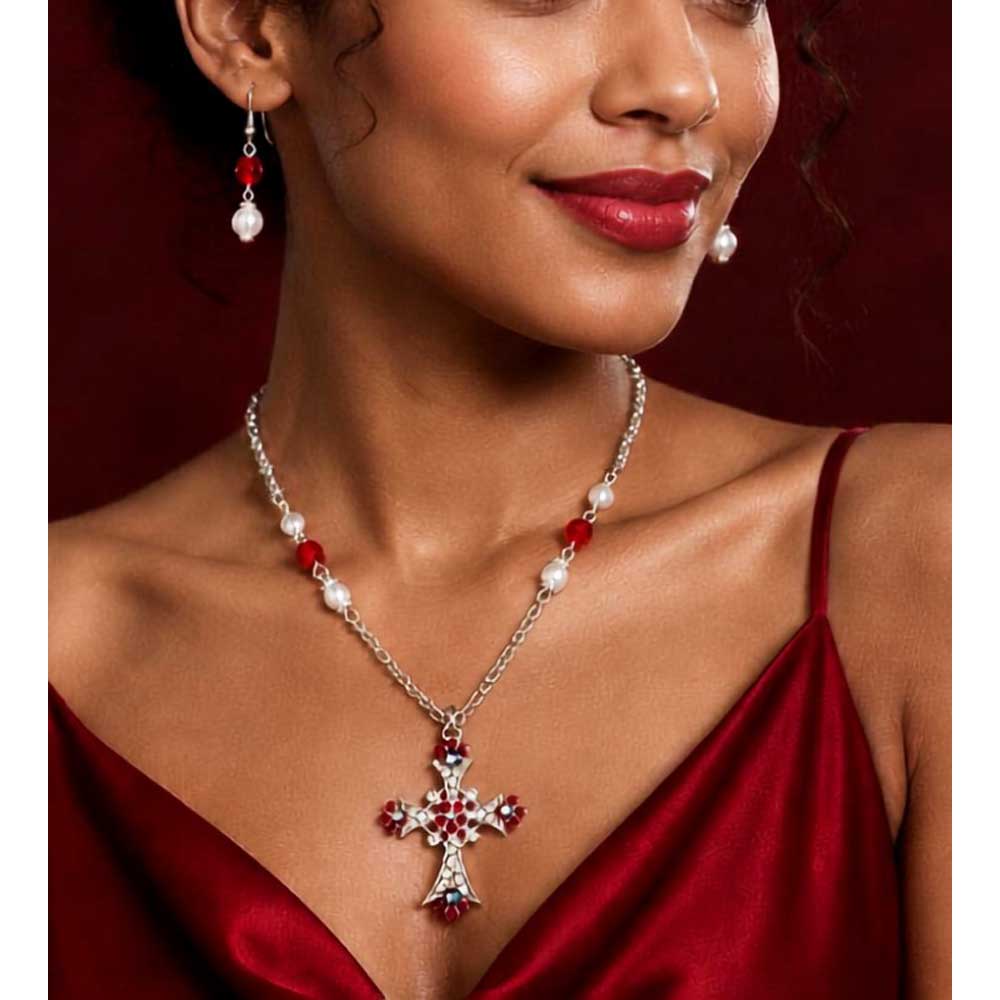 A close-up of a woman wearing a silver cross pendant necklace with red and white beads, matching drop earrings, and a deep red satin dress against a dark red background. Lilylin Designs