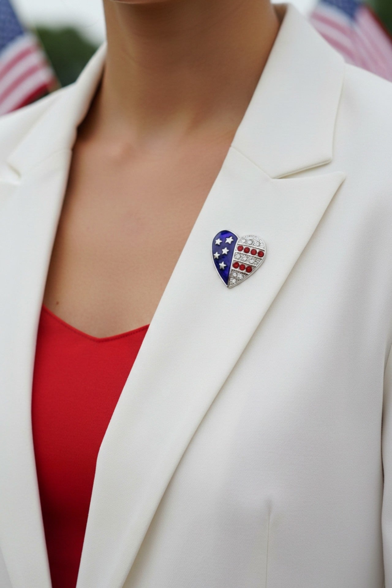 Model wearing silver small patriotic heart brooch pin on a white jacket with a red top and blurred American flags in the background - Lilylin Designs