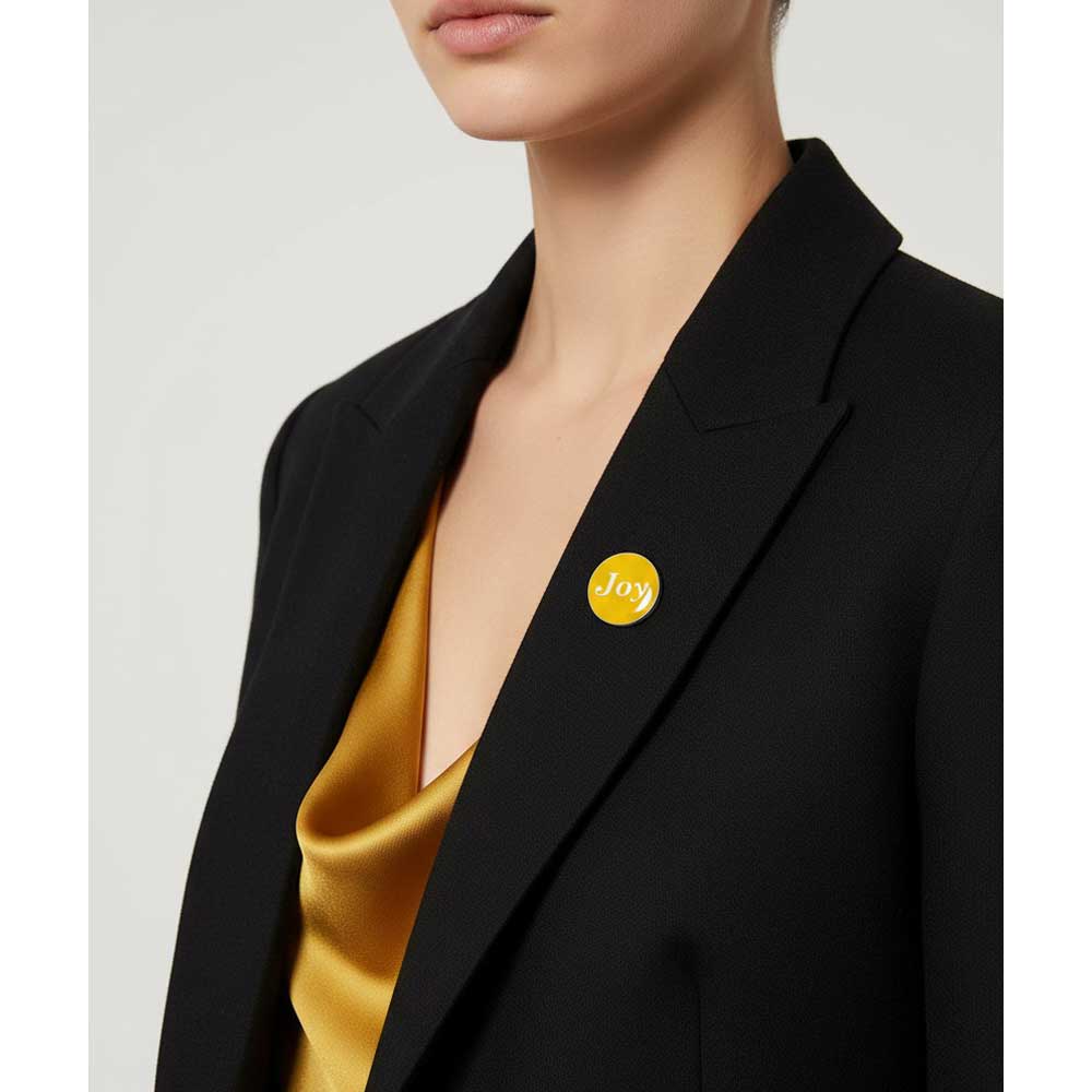 A close-up of a model wearing a black blazer over a gold satin top, featuring a small round yellow pin on the lapel with the word 'Joy' written in white script. Lilylin Designs