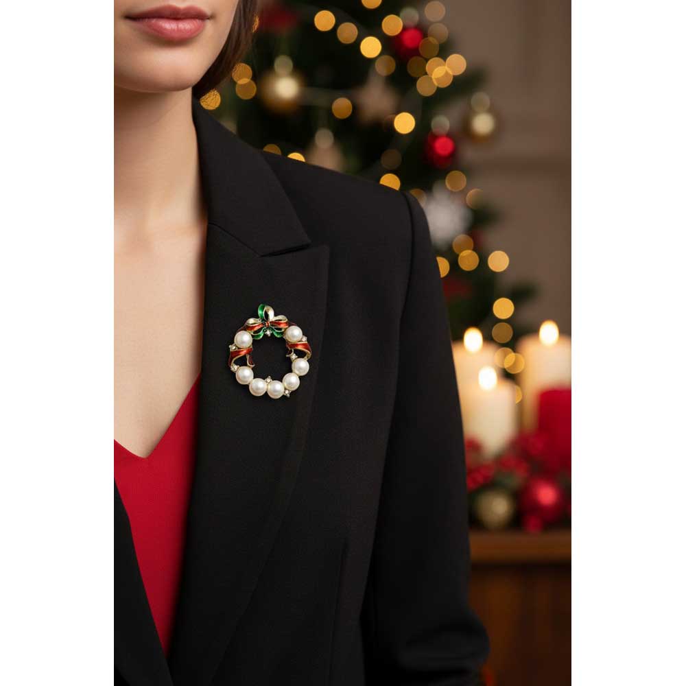 A close-up of a woman wearing a black blazer over a red top, featuring a holiday wreath brooch with white pearls, green leaves, and red and gold accents on the left lapel, with a blurred Christmas tree in the background. Lilylin Designs