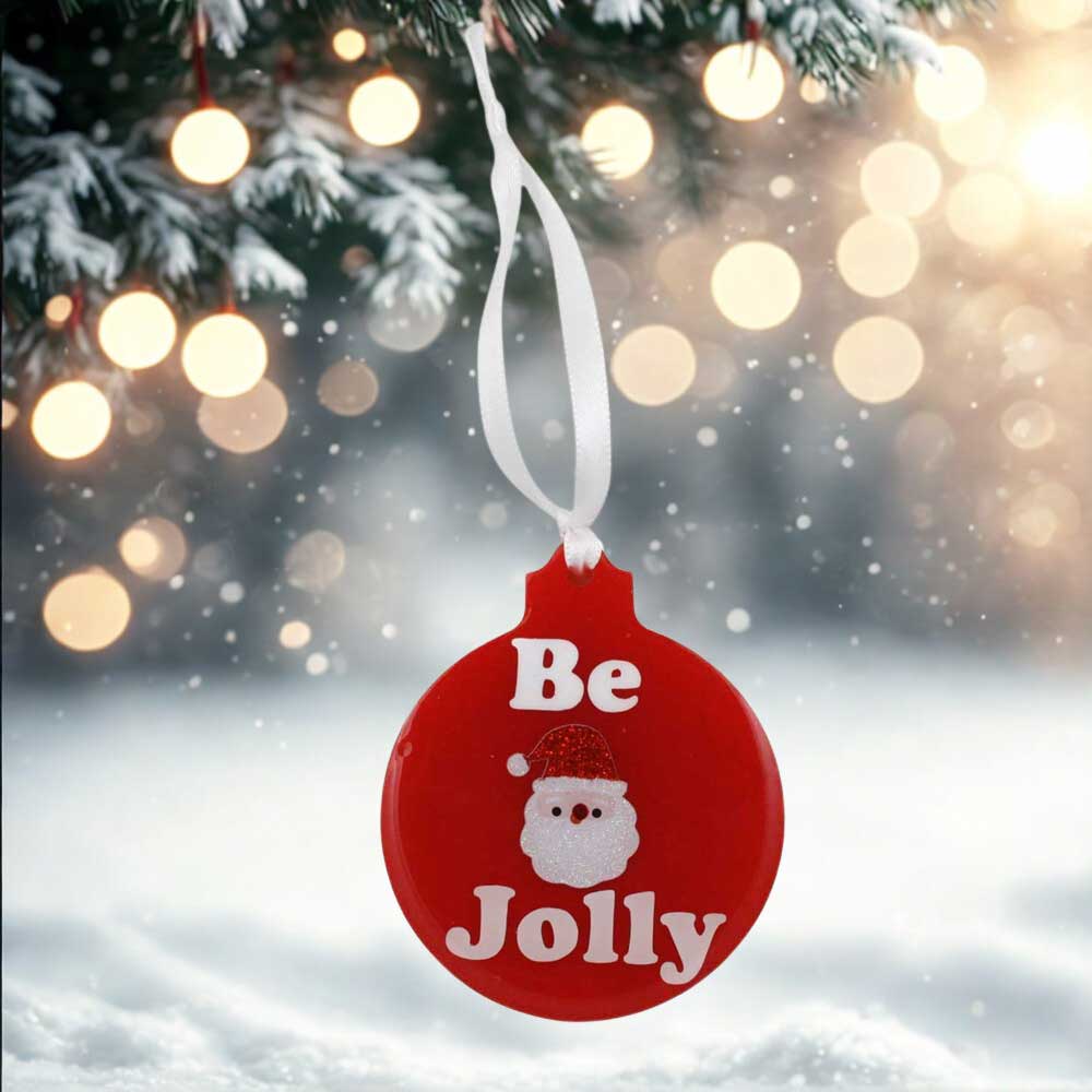 Red Christmas ornament with 'Be Jolly' text and Santa Claus face, hanging from a tree with lights and snow in the background. | Lilylin Designs