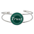 A silver-toned metal cuff bracelet featuring a round green disc with the word 'Trust' printed in white script, shown on a plain white background – Lilylin Designs