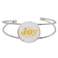 A silver-toned metal cuff bracelet featuring a round white disc with the word 'Joy' printed in yellow script, shown on a plain white background – Lilylin Designs