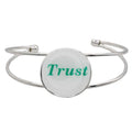 A silver-toned metal cuff bracelet featuring a round white disc with the word "Trust" printed in green script, shown on a plain white background – Lilylin Designs